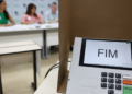 Urna eletrônica em seção eleitoral com integrantes da mesa receptora de votos ao fundo.