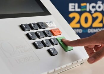 Imagem ilustrativa: dedo pressiona a tecla verde de confirmação do teclado de uma urna eletrônica, ao lado da marca das Eleições 2026