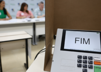 Urna eletrônica em seção eleitoral com integrantes da mesa receptora de votos ao fundo.