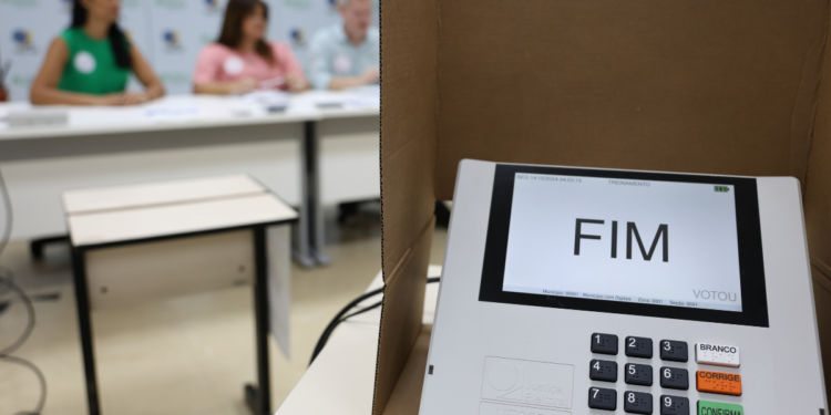 Urna eletrônica em seção eleitoral com integrantes da mesa receptora de votos ao fundo.