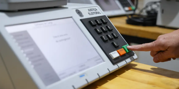Dedo pressionando a tecla verde de confirmação de uma urna eletrônica da Justiça Eleitoral, com outros equipamentos enfileirados sobre a bancada ao fundo.