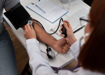 Imagem ilustrativa de profissional de jaleco branco aferindo a pressão arterial de um paciente em consultório, com prancheta e computador sobre a mesa.