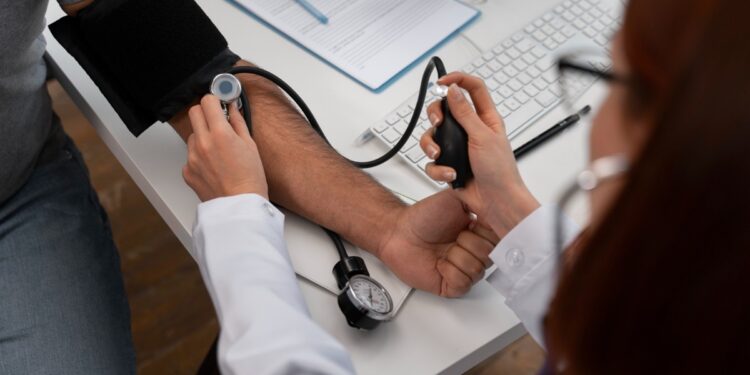 Imagem ilustrativa de profissional de jaleco branco aferindo a pressão arterial de um paciente em consultório, com prancheta e computador sobre a mesa.