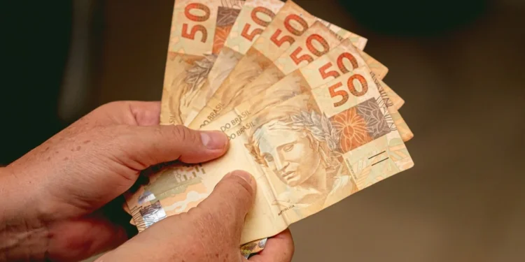 Mãos seguram cédulas de real, representando o pagamento do INSS de até R$ 8.475,55 para beneficiários com cartão final 5 e 0.