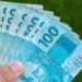 Cédulas de R$ 100 sendo seguradas representam o Saque Calamidade do FGTS de até R$ 6.220 para moradores de cidades recentemente habilitadas.