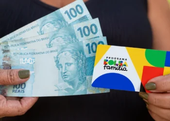 Mãos segurando notas de 100 reais e cartão do Bolsa Família representando o novo valor mínimo de R$ 691