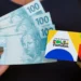 Mãos segurando notas de 100 reais e cartão do Bolsa Família representando o novo valor mínimo de R$ 691