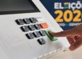 Dedo pressionando a tecla verde de confirmação da urna eletrônica ao lado da marca Eleições 2026