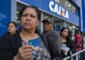 Fila de brasileiros em frente a uma agência da Caixa durante paralisação, com cliente segurando cartão de benefício