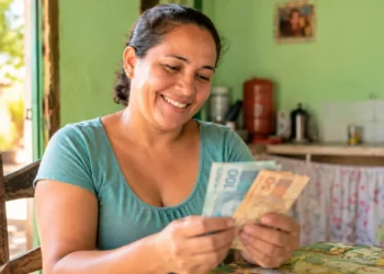 Mulher segura dinheiro em casa, representando o pagamento de R$ 1.200 do programa Mães de Pernambuco.