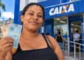 Mulher segura notas de real em frente a uma agência da Caixa representando o pagamento de R$ 300 do programa Mães de Pernambuco