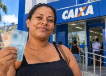 Mulher segura notas de real em frente a uma agência da Caixa representando o pagamento de R$ 300 do programa Mães de Pernambuco