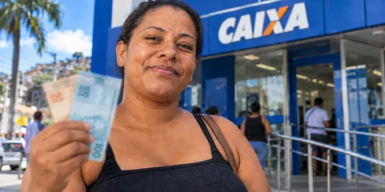 Mulher segura notas de real em frente a uma agência da Caixa representando o pagamento de R$ 300 do programa Mães de Pernambuco