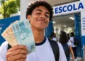 Estudante segura notas de reais na entrada de escola durante pagamento do programa Pé-de-Meia