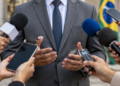 Porta-voz de político durante entrevista com jornalistas utilizando microfones e celulares.