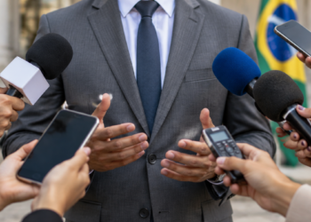 Porta-voz de político durante entrevista com jornalistas utilizando microfones e celulares.