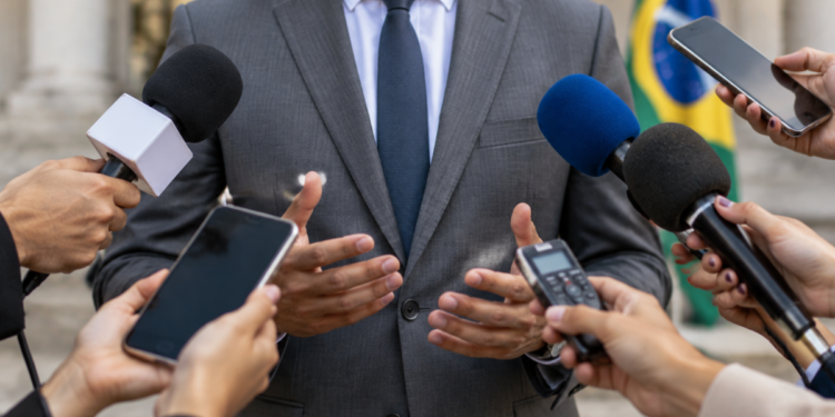 Porta-voz de político durante entrevista com jornalistas utilizando microfones e celulares.