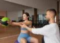 Profissional de educação física orienta mulher durante exercício com bola em academia.