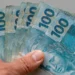 Mão segura cédulas de R$ 100 em referência aos recursos previstos no Projeto de Lei Orçamentária de 2027.