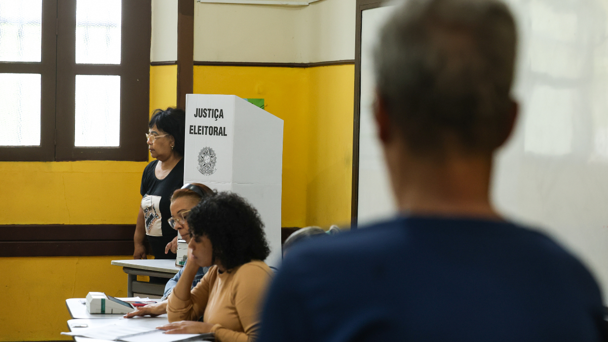 Eleitor em seção eleitoral com urna eletrônica e integrantes da mesa receptora de votos durante as Eleições 2026.