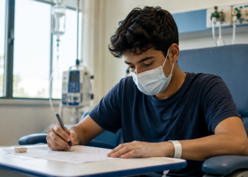 Estudante usa máscara e realiza prova escrita em ambiente hospitalar durante tratamento de saúde.