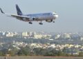 Avião comercial em aproximação final para pouso, sobrevoando a área urbana próxima ao aeroporto em dia de céu claro.