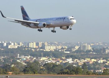 Avião comercial em aproximação final para pouso, sobrevoando a área urbana próxima ao aeroporto em dia de céu claro.