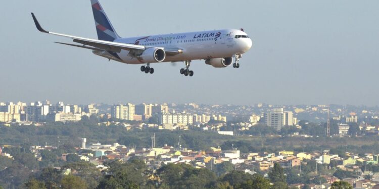 Avião comercial em aproximação final para pouso, sobrevoando a área urbana próxima ao aeroporto em dia de céu claro.