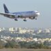 Avião comercial em aproximação final para pouso, sobrevoando a área urbana próxima ao aeroporto em dia de céu claro.