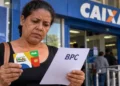 Mulher segura cartão do Bolsa Família e documento sobre solicitação do BPC em frente a uma agência da Caixa
