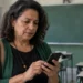 Professora consultando o celular em sala de aula, representando a orientação do MEC para verificar o local de prova com antecedência.