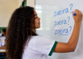 Estudante escrevendo em quadro branco as palavras zueira, zuera e zoeira durante aula de português.