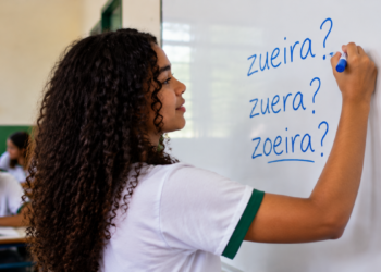 Estudante escrevendo em quadro branco as palavras zueira, zuera e zoeira durante aula de português.