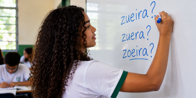 Estudante escrevendo em quadro branco as palavras zueira, zuera e zoeira durante aula de português.